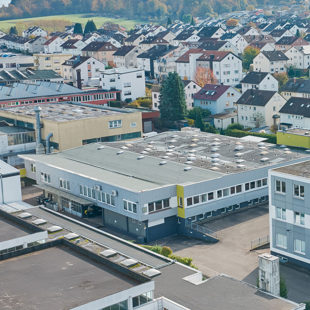 A large industrial building with a flat roof is surrounded by residential houses and greenery in a suburban area on a clear day.