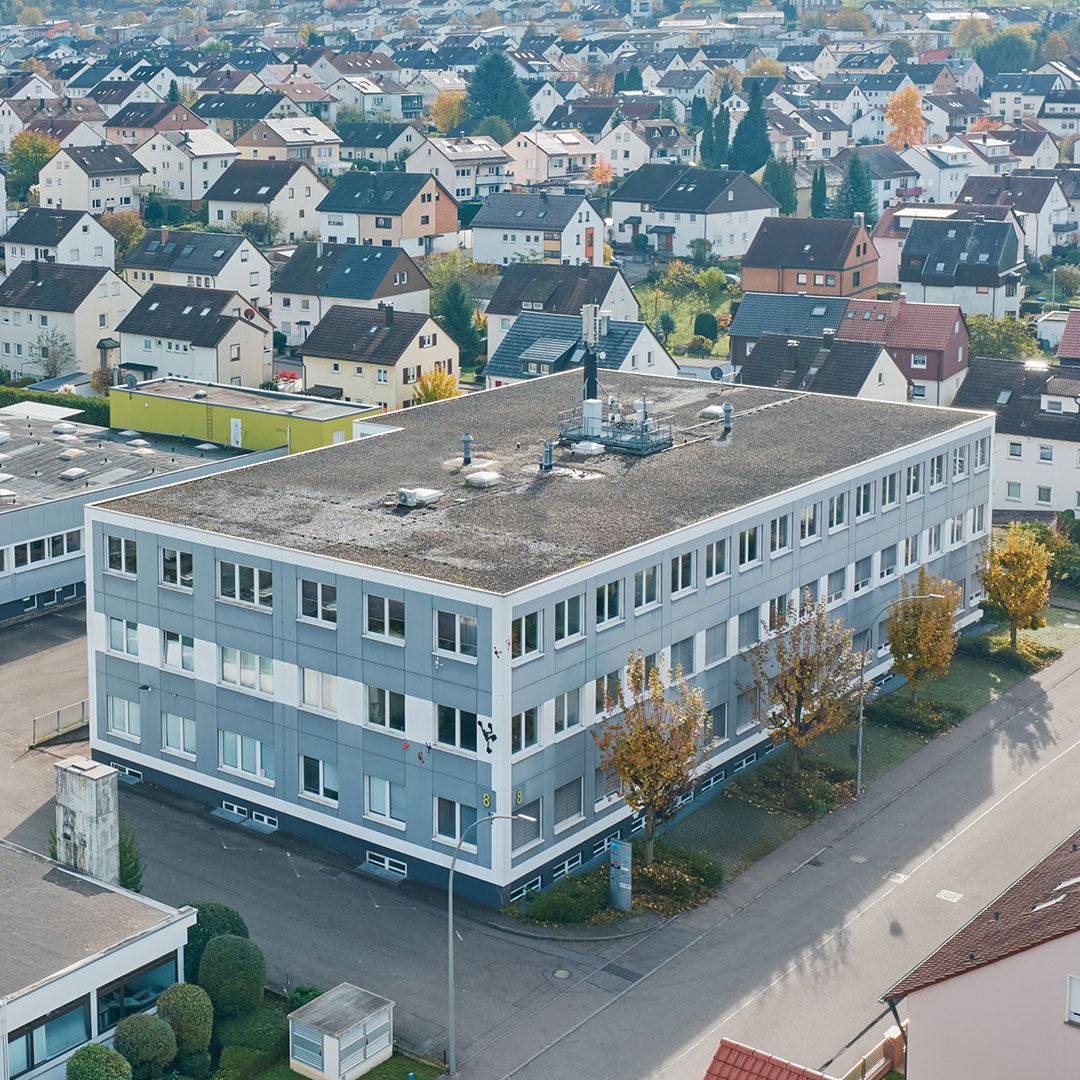 Aerial view of a rectangular, three-story building with a flat roof, surrounded by trees and streets, in a residential neighborhood with many houses.