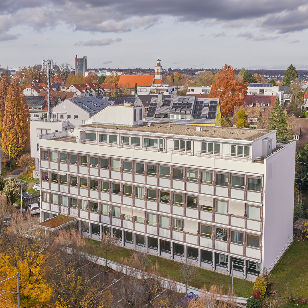 A four-story office building with large windows, surrounded by autumn trees and residential houses, under a cloudy sky. Solar panels are visible on nearby rooftops.