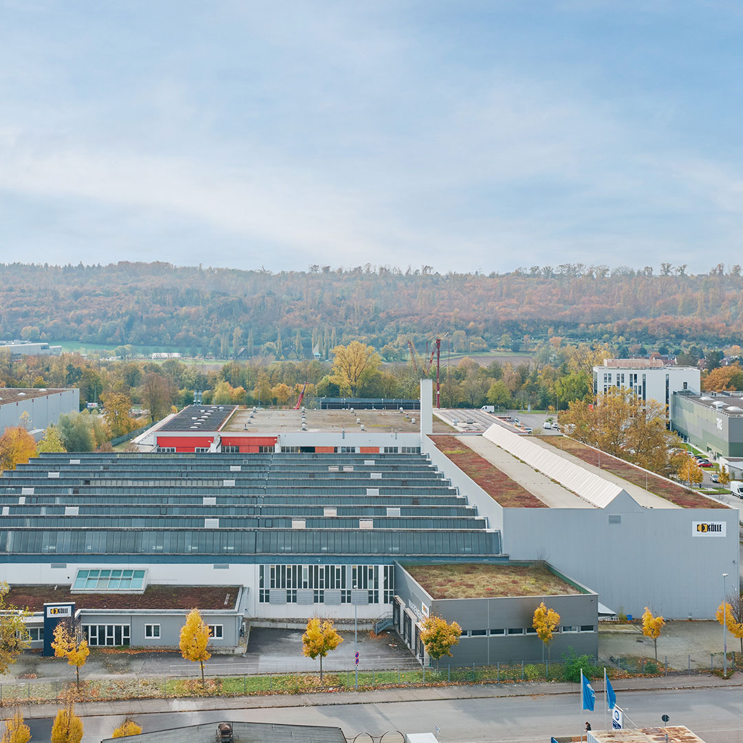 Aerial view of an industrial complex with multiple warehouse buildings, surrounded by trees with autumn foliage and hills in the background under a clear sky.