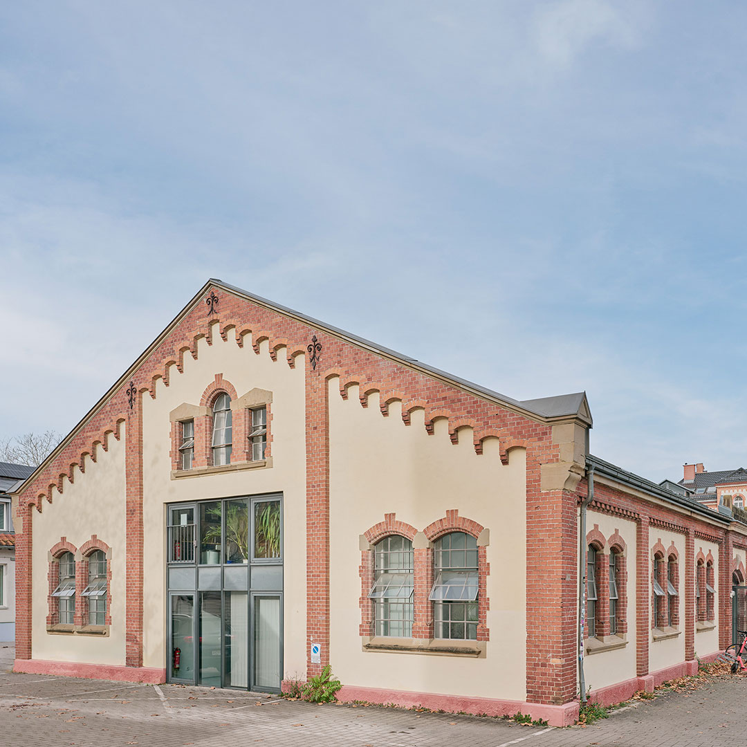 A historic, brick industrial building with arched windows and a modern glass entrance, set against a clear sky. Some greenery is visible through the windows.