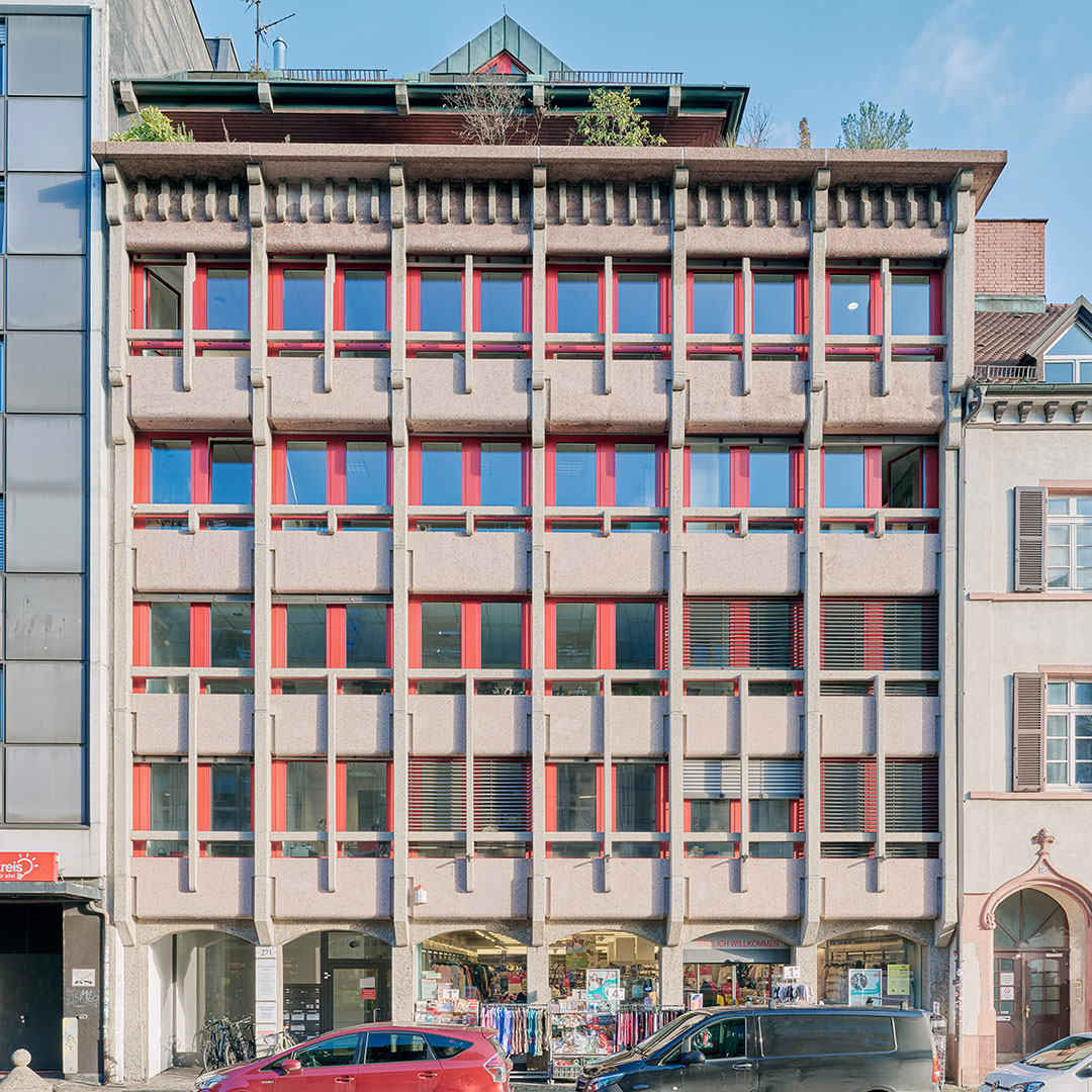 A modern building with red-framed windows, concrete facade, and ground-level shops displaying clothes and goods on racks; cars are parked in front on the street.