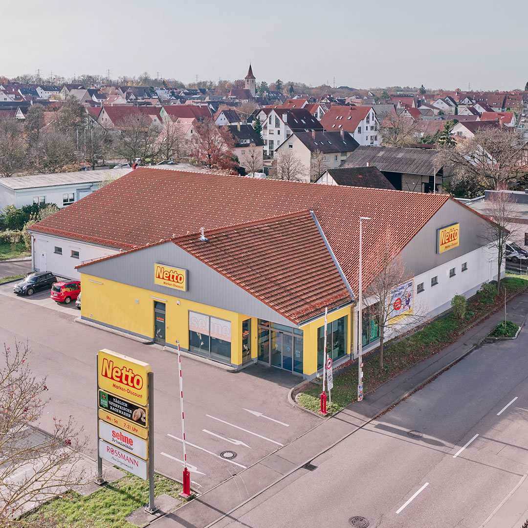 A Netto supermarket with a red roof and yellow exterior sits at a street corner in a suburban area with houses and trees in the background. A sign for the store stands near the entrance.