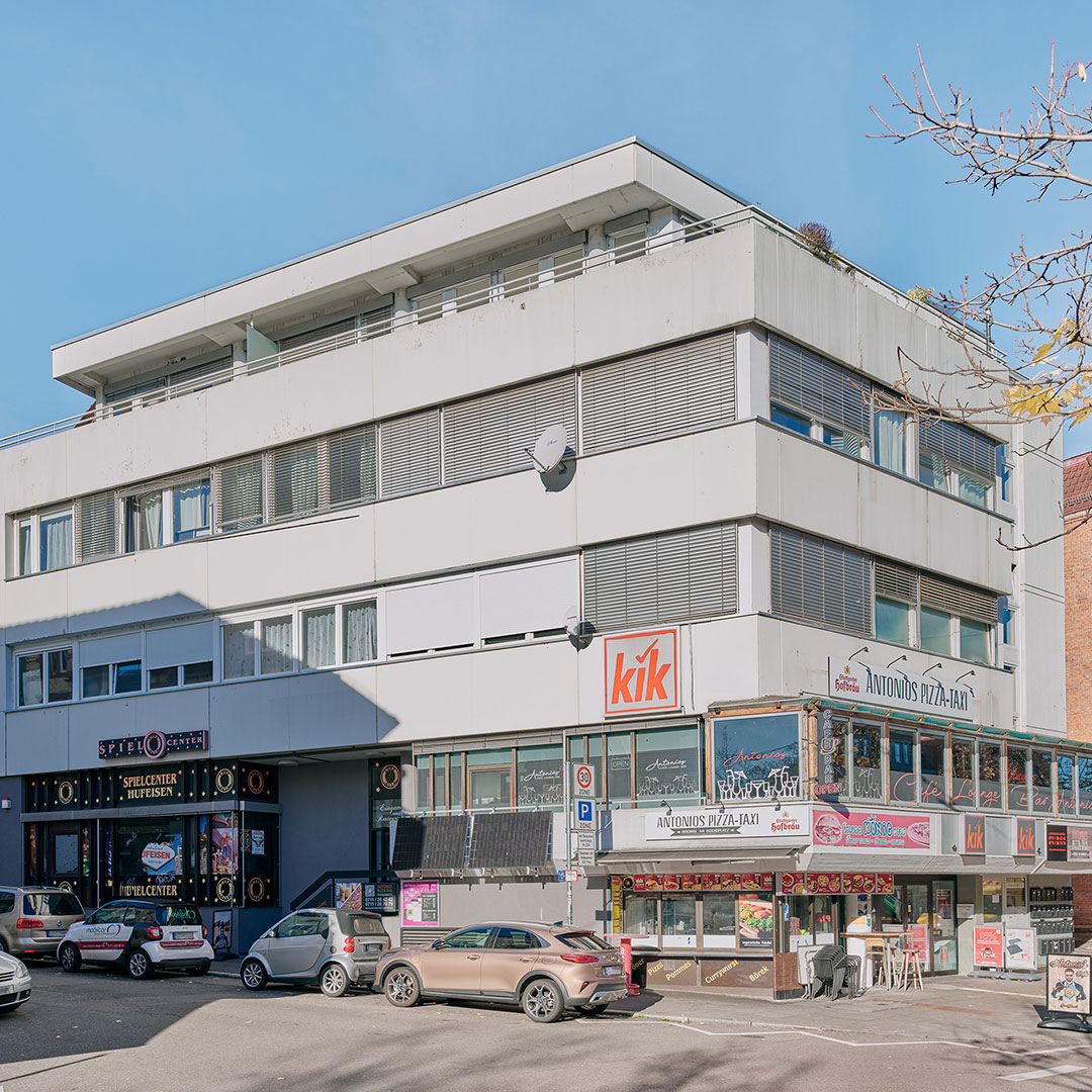 A white, modern commercial building with shops and restaurants on the ground floor, including kik and Antons Pizza-Bar, with parked cars in front and a clear, blue sky above.
