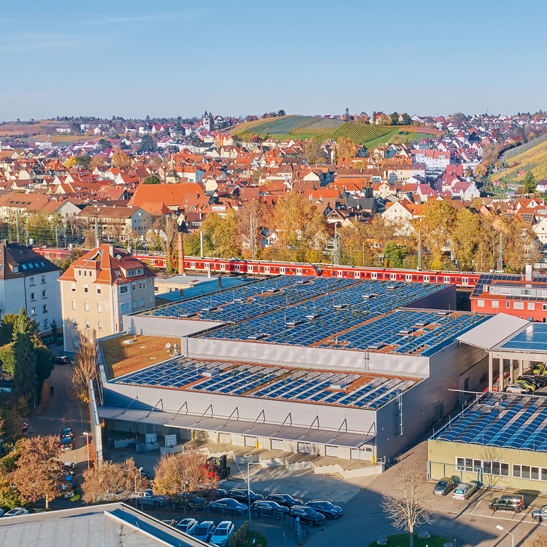 Aerial view of a town with red-roofed buildings, a train passing by, and a modern building in the foreground covered in solar panels. Autumn trees and rolling hills are visible in the background.