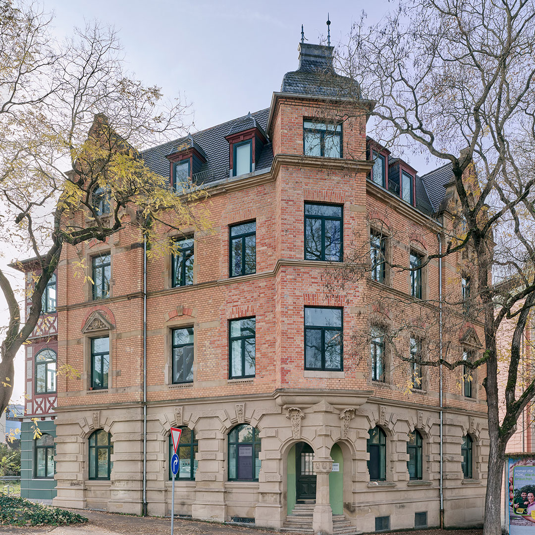 A historic three-story brick building with large windows, arched doorways, and decorative trim stands at a street corner, surrounded by leafless trees and a traffic sign.