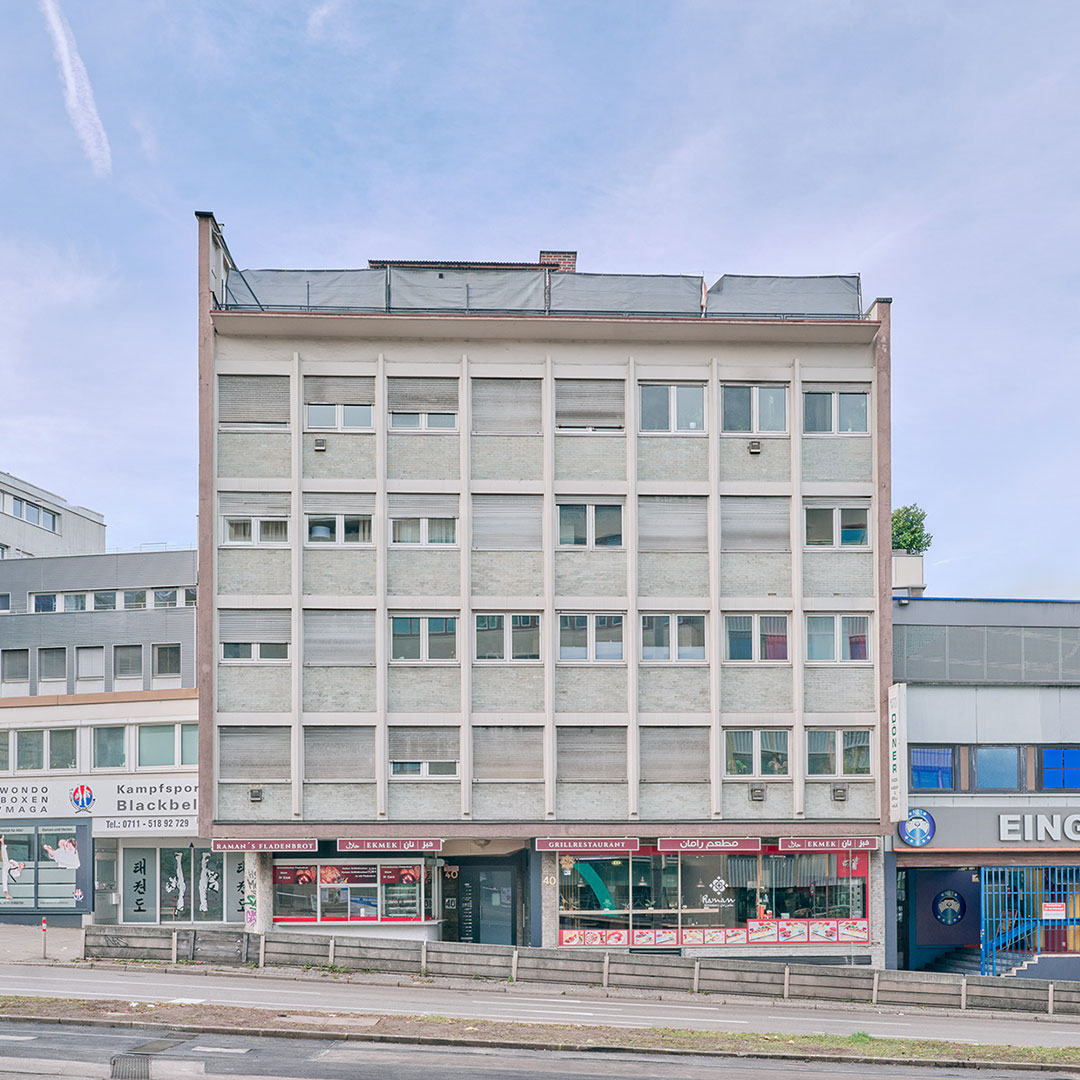 A gray, five-story rectangular building with small windows and shops on the ground floor stands on a city street; neighboring buildings are partially visible.