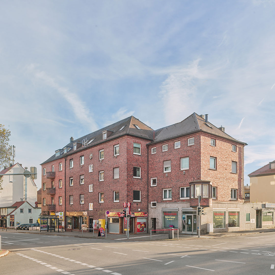 A red brick building with multiple shops at street level stands at a corner under a clear sky. Several windows and doors are visible, and cars are parked along the street.