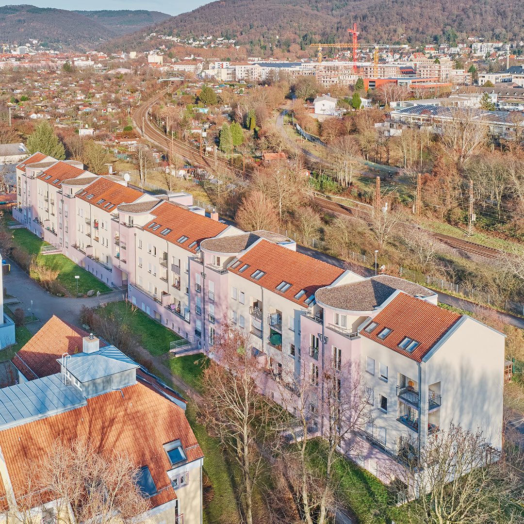 Aerial view of pink apartment buildings with red roofs near railway tracks, trees, and a distant town surrounded by hills under a clear sky.