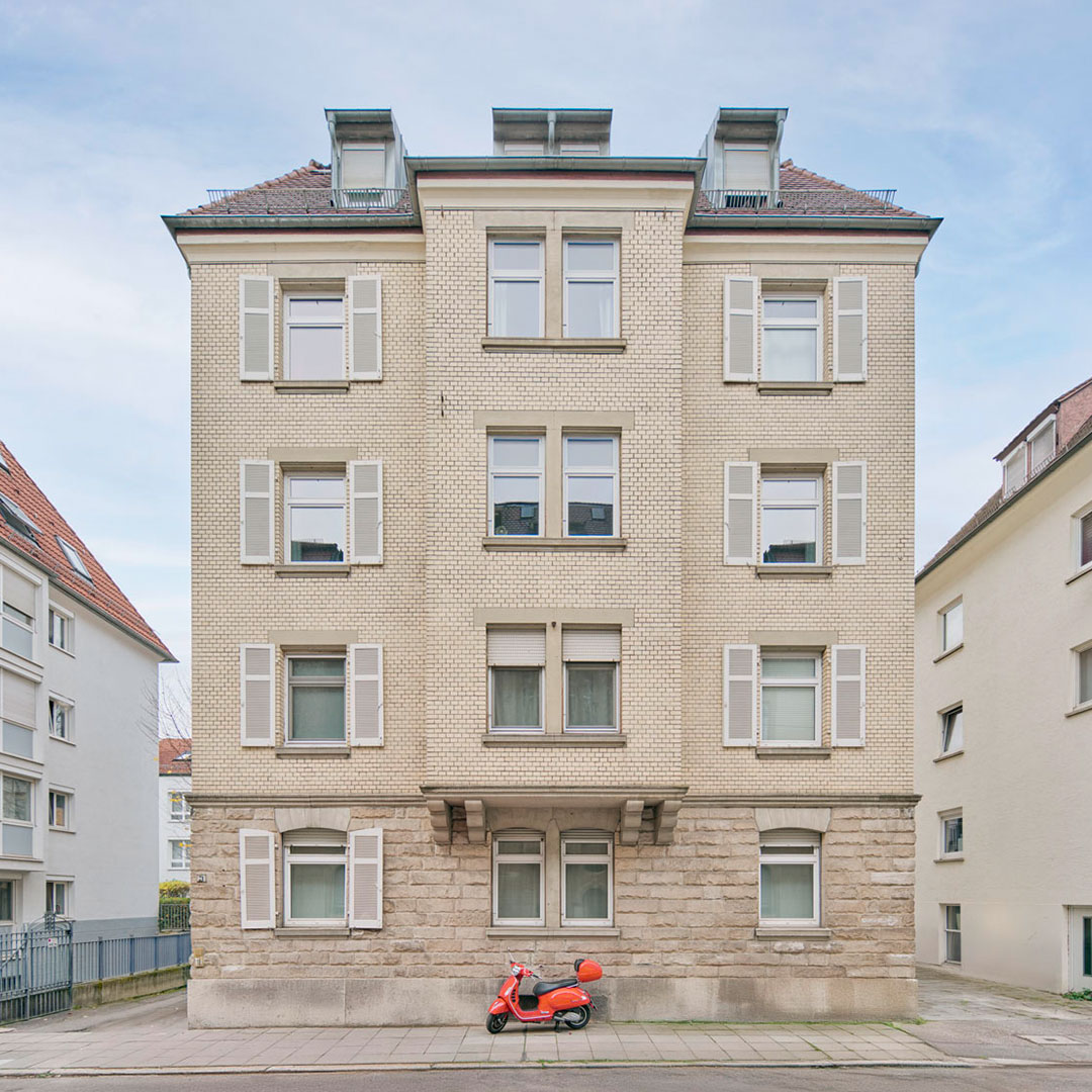 A beige apartment building with symmetrical windows and shutters, and a red scooter with a helmet parked in front on the sidewalk.