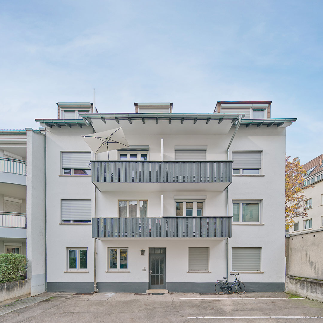 A three-story white apartment building with gray balconies and closed shutters. A bicycle is parked by the entrance, and an umbrella stands on the top balcony. The sky is clear with a few clouds.