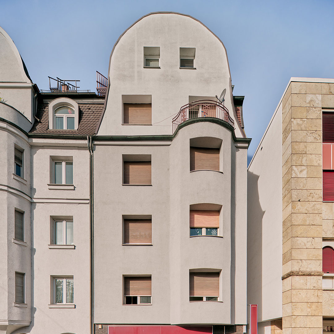 A white, multi-story building with rounded roof edges and several windows with closed shades, flanked by neighboring buildings under a clear blue sky.
