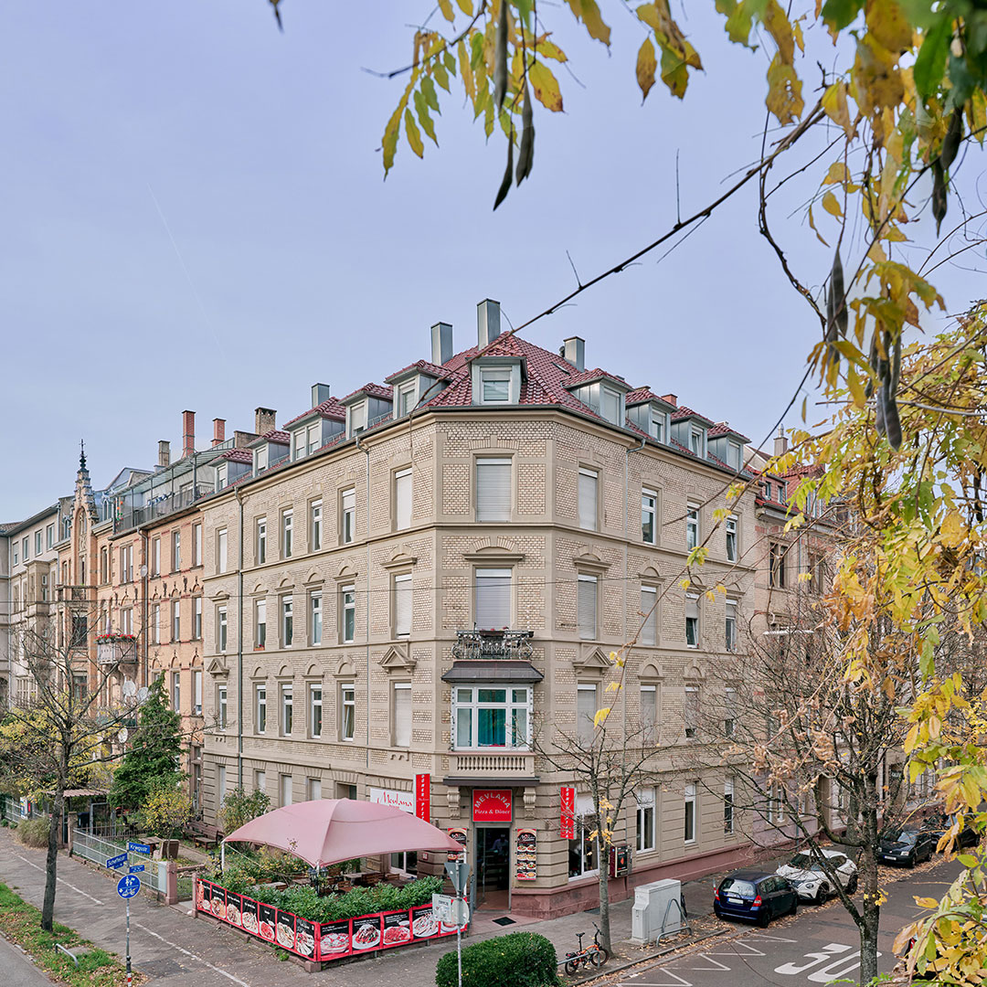 A beige, four-story corner building with a red-roof and a restaurant on the ground floor, outdoor seating under pink umbrellas, and autumn trees lining the street.