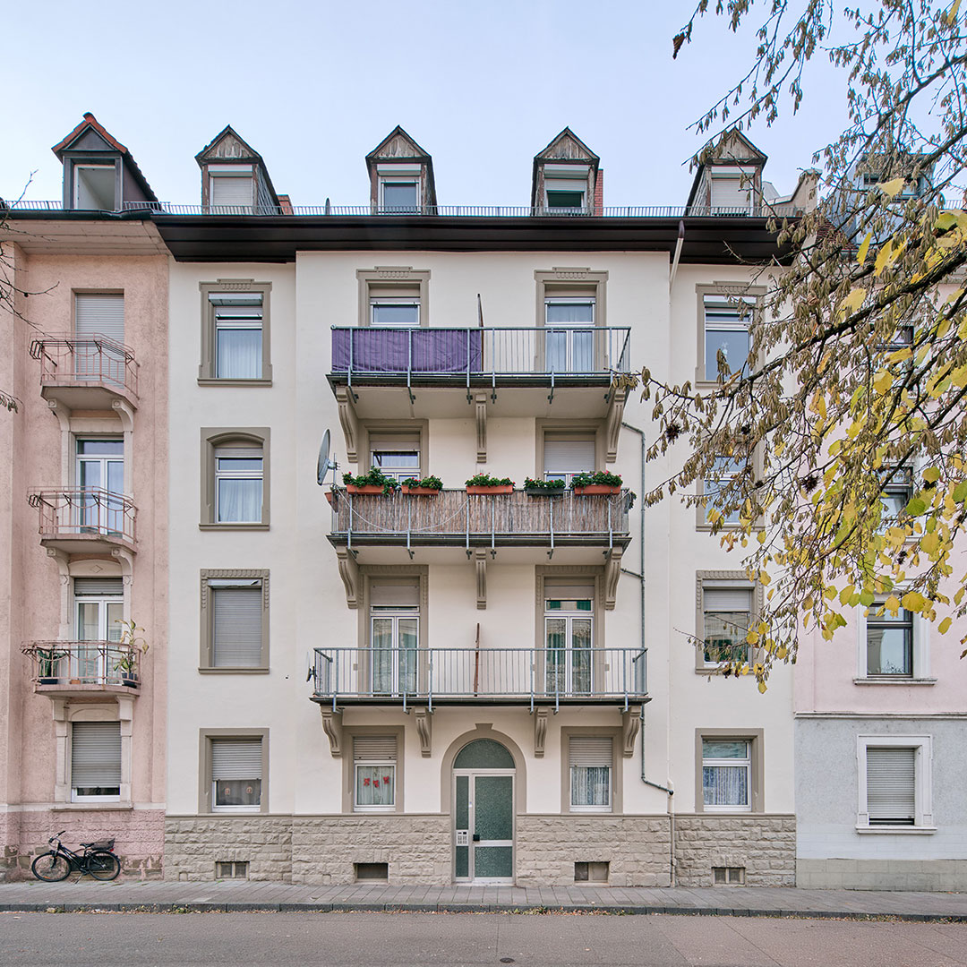 A four-story apartment building with balconies, potted plants, and a glass entrance door stands between two similar buildings. A bicycle is parked on the sidewalk, and tree branches frame the right side of the image.