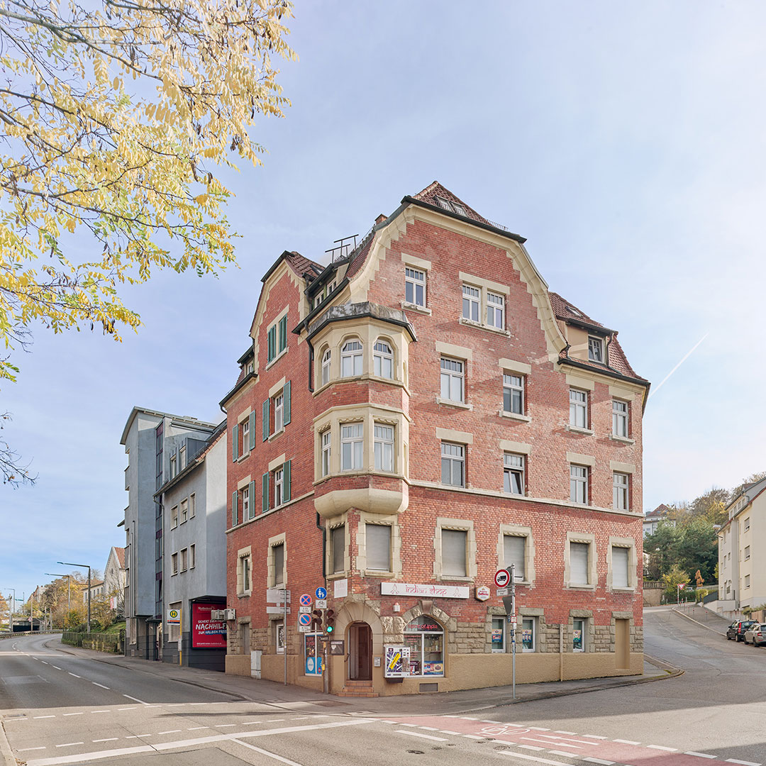 A four-story red brick building with white trim stands on a street corner. There are street signs, a few shops on the ground floor, and a tree with autumn leaves on the left. The sky is clear and blue.
