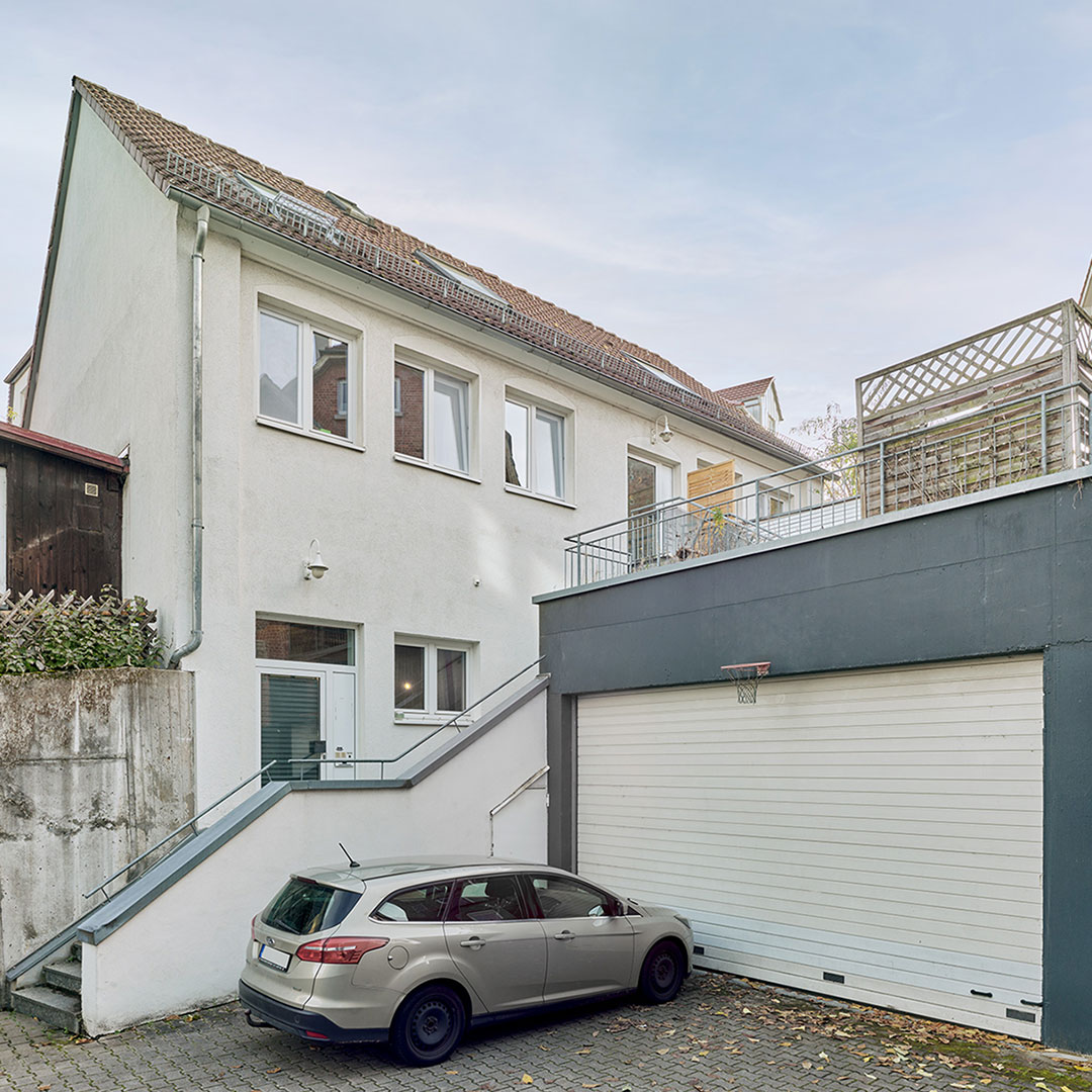 A white two-story house with a sloped roof, an outdoor staircase, and a gray garage. A silver car is parked in front of the garage door. Some leaves are scattered on the ground.
