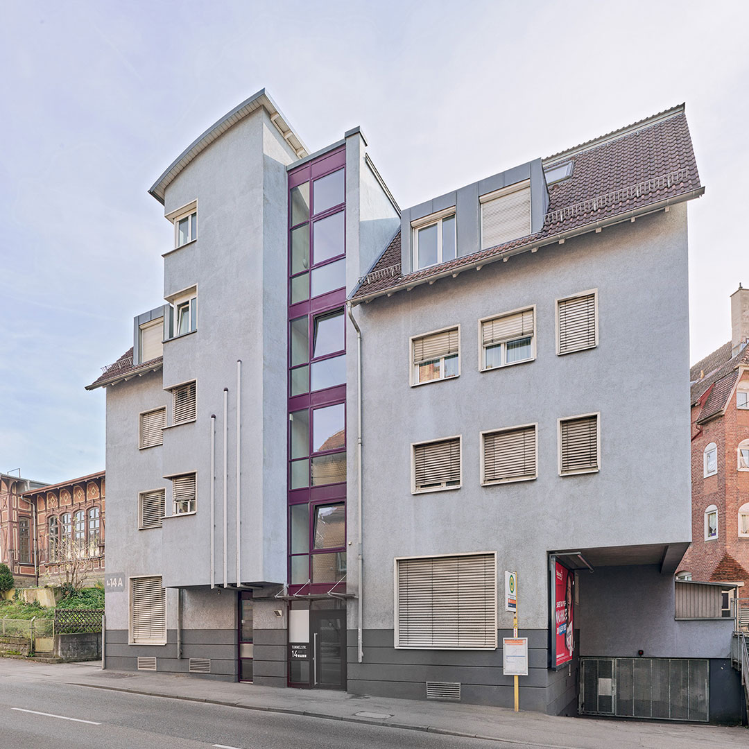 A modern, light-gray, multi-story building with purple-framed glass stairwell, large windows with shutters, and a covered parking space on a quiet street.
