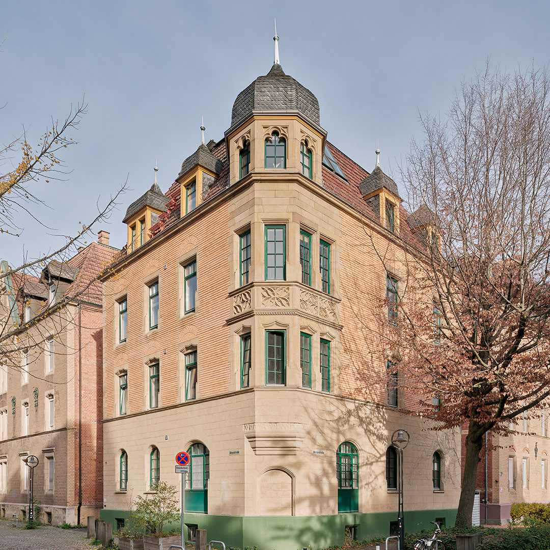 A historic three-story corner building with tan brick, green-framed windows, and a turret-like roof feature. Leafless trees line the sidewalk and a bike is parked out front.