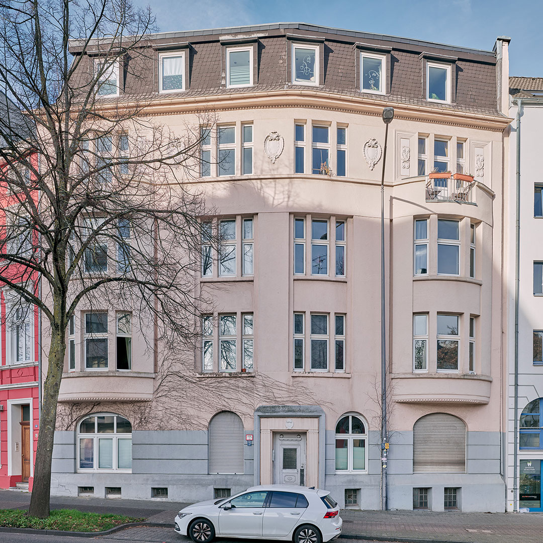 A beige, four-story apartment building with curved facade and decorative details, tall windows, and a white car parked in front. A tree with bare branches stands on the sidewalk.