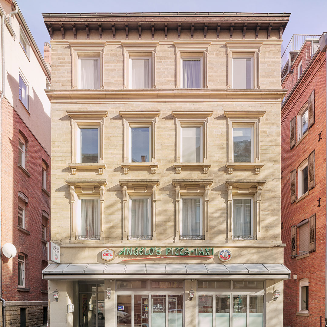 Four-story beige building with tall windows, decorative trim, and a pizza restaurant named Angelos Pizza Taxi on the ground floor. Red brick buildings are on either side. Sunlight brightens the facade.