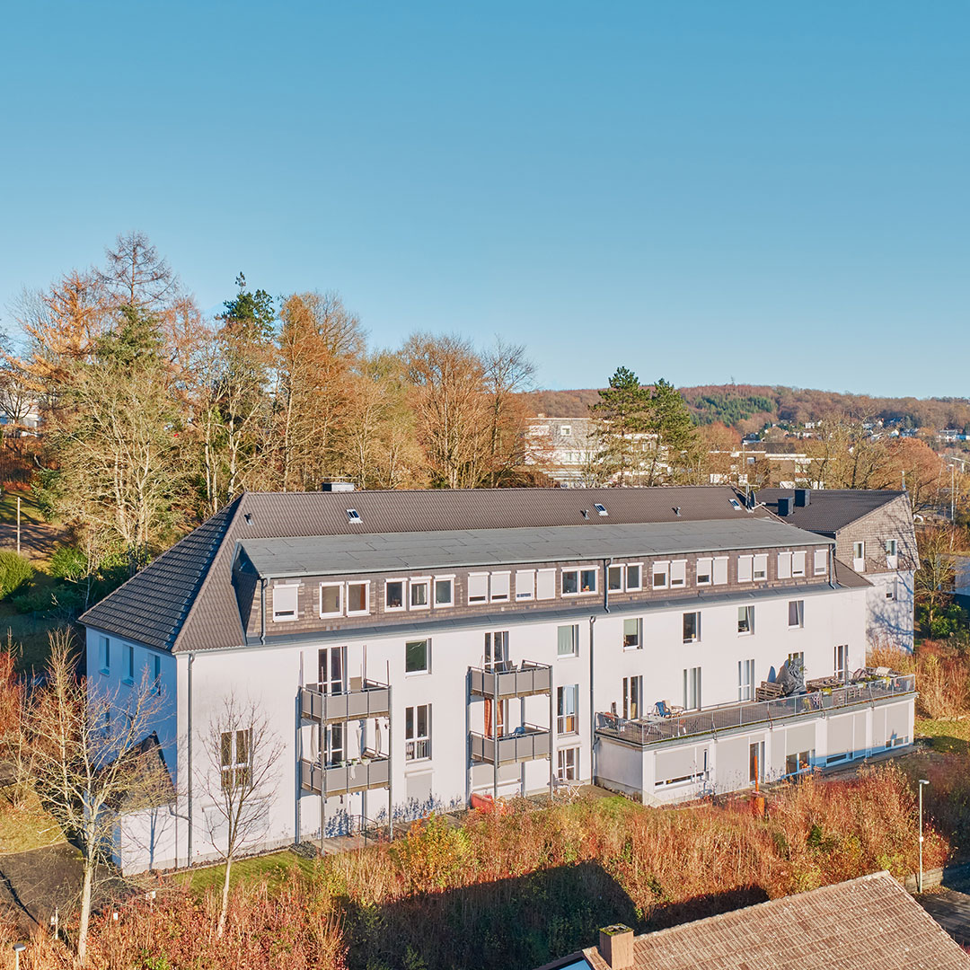 A modern, light-colored apartment building with balconies, set among autumn trees and bushes, under a clear blue sky. Other buildings and forested hills are visible in the background.