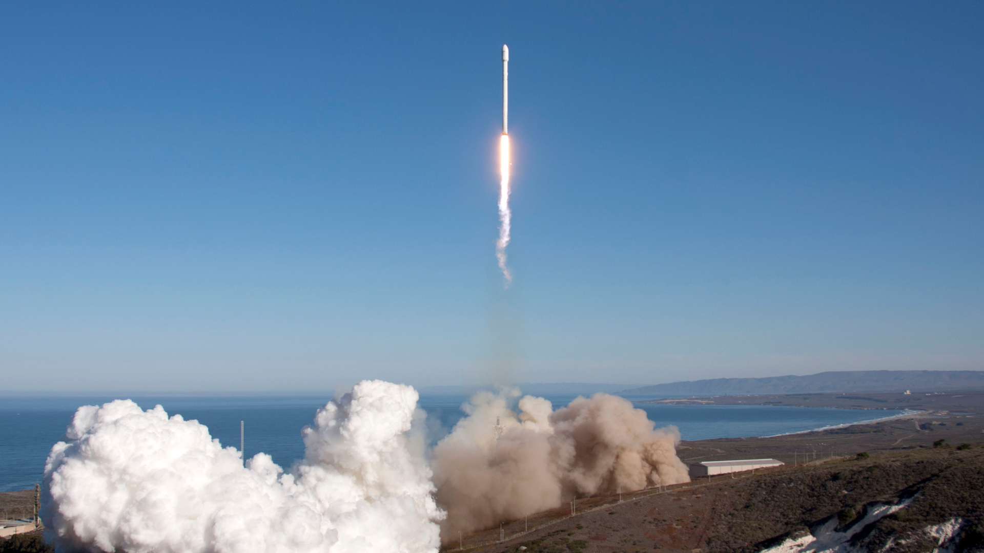Eine weiße Rakete schießt senkrecht in den strahlend blauen Himmel über einer Küstenlandschaft und hinterlässt am Startort eine große Rauchwolke.